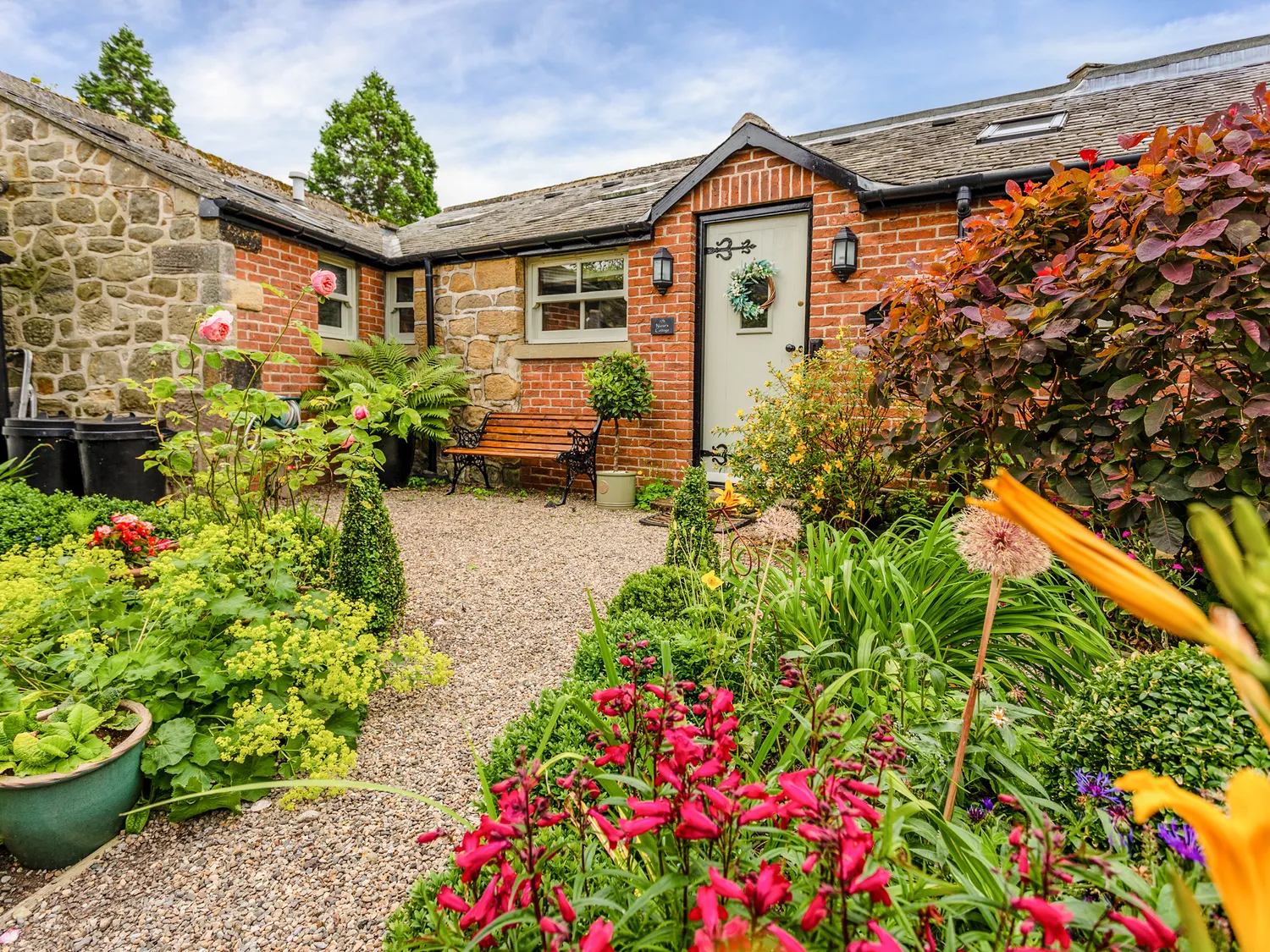 A quaint stone and brick house with a gable roof, surrounded by a lush, colourful garden featuring a variety of flowers and plants. A gravel path leads to the front door.