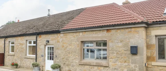 Stone cottage with two types of tiled roofs, white window frames, and a white front door flanked by two potted plants.