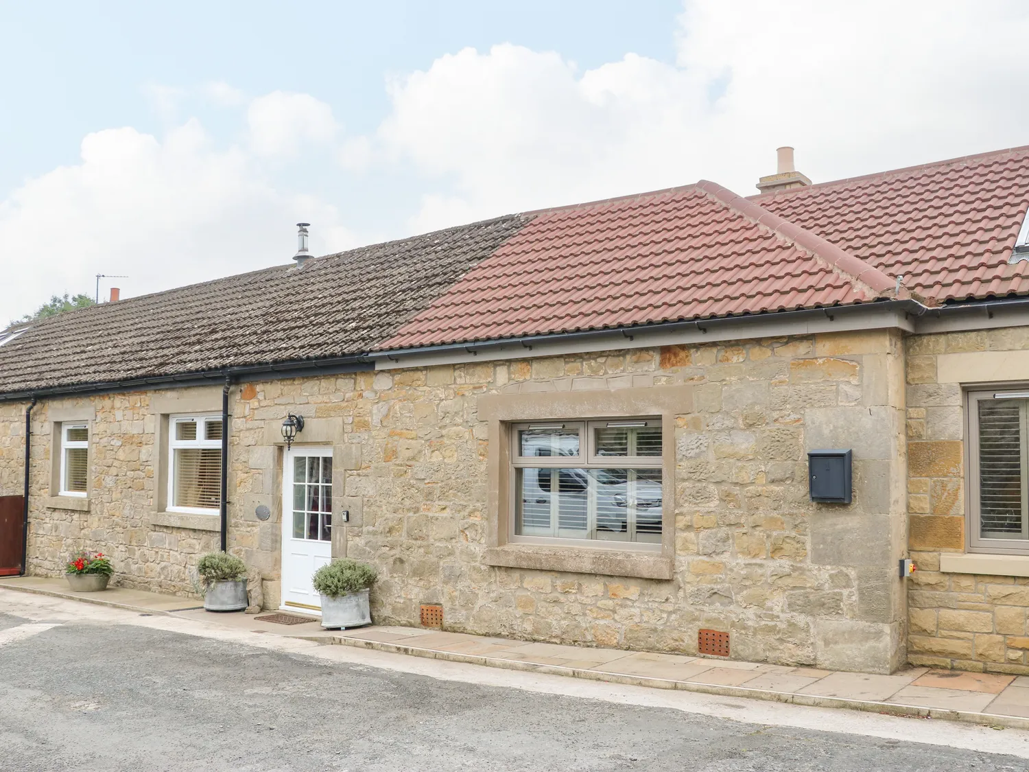 Stone cottage with two types of tiled roofs, white window frames, and a white front door flanked by two potted plants.