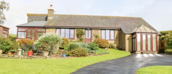 Single-storey house with a conservatory, stone façade, and red-framed windows, surrounded by well-manicured gardens and a curved asphalt driveway.