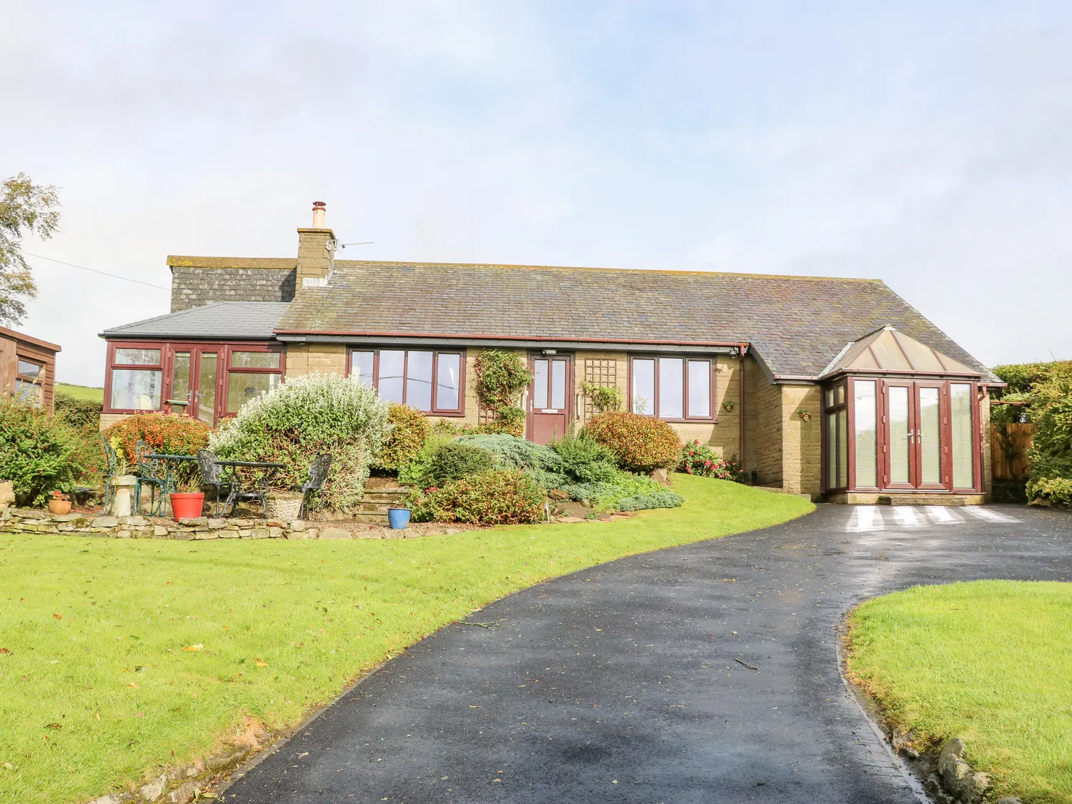 Single-storey house with a conservatory, stone façade, and red-framed windows, surrounded by well-manicured gardens and a curved asphalt driveway.