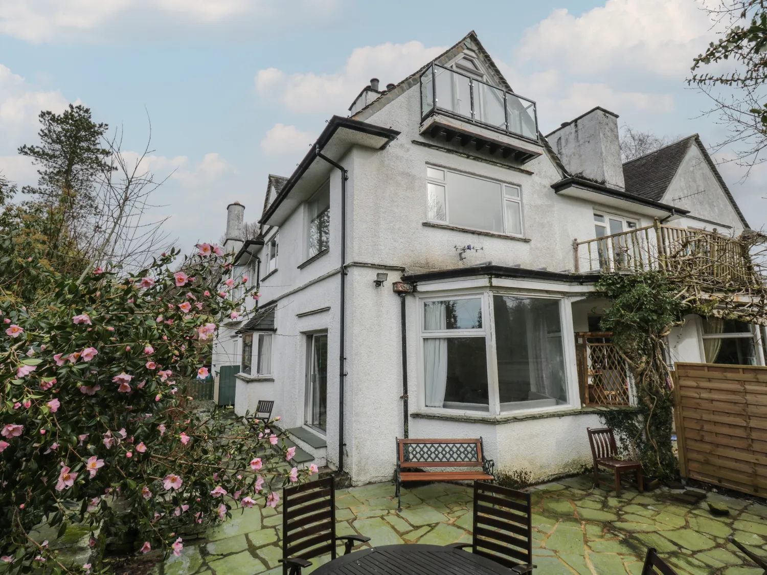 Exterior view of a white multi-storey house with a patio area, surrounded by a garden with blooming pink flowers.