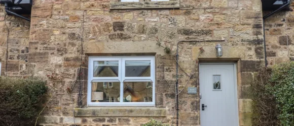 Stone cottage facade with a grey door, a window to the left of the door, and a smaller window above.