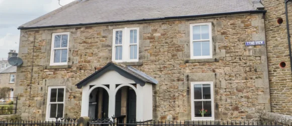 Stone-built house with four windows, a central arched porch, a grey roof, and iron fencing. Street sign "Tyne View" is visible on the right side of the house.