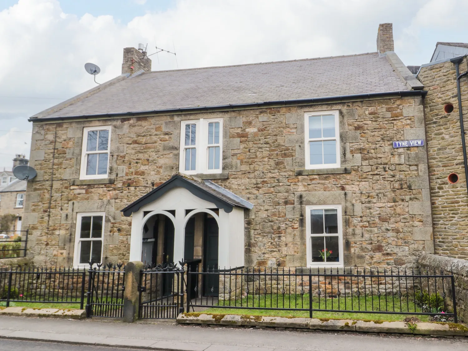 Stone-built house with four windows, a central arched porch, a grey roof, and iron fencing. Street sign "Tyne View" is visible on the right side of the house.