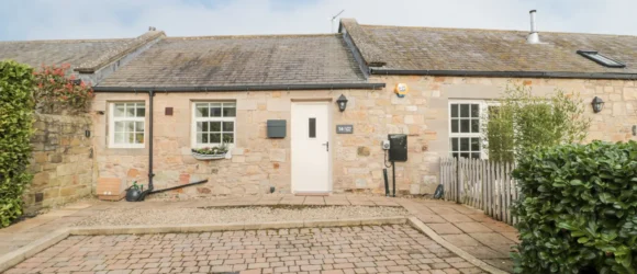 Front view of a stone cottage with a tiled roof, white door, and white-framed windows. There's a small front garden with a potted plant and a cobblestone driveway.