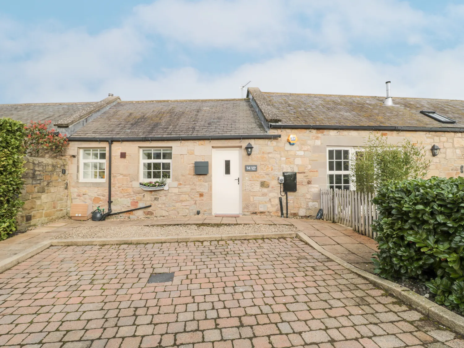 Front view of a stone cottage with a tiled roof, white door, and white-framed windows. There's a small front garden with a potted plant and a cobblestone driveway.