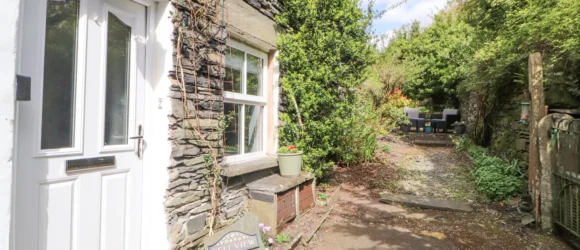 Front entrance of a rustic stone cottage with a sign reading "Fell View," adjacent to a path leading to a garden area with outdoor seating.