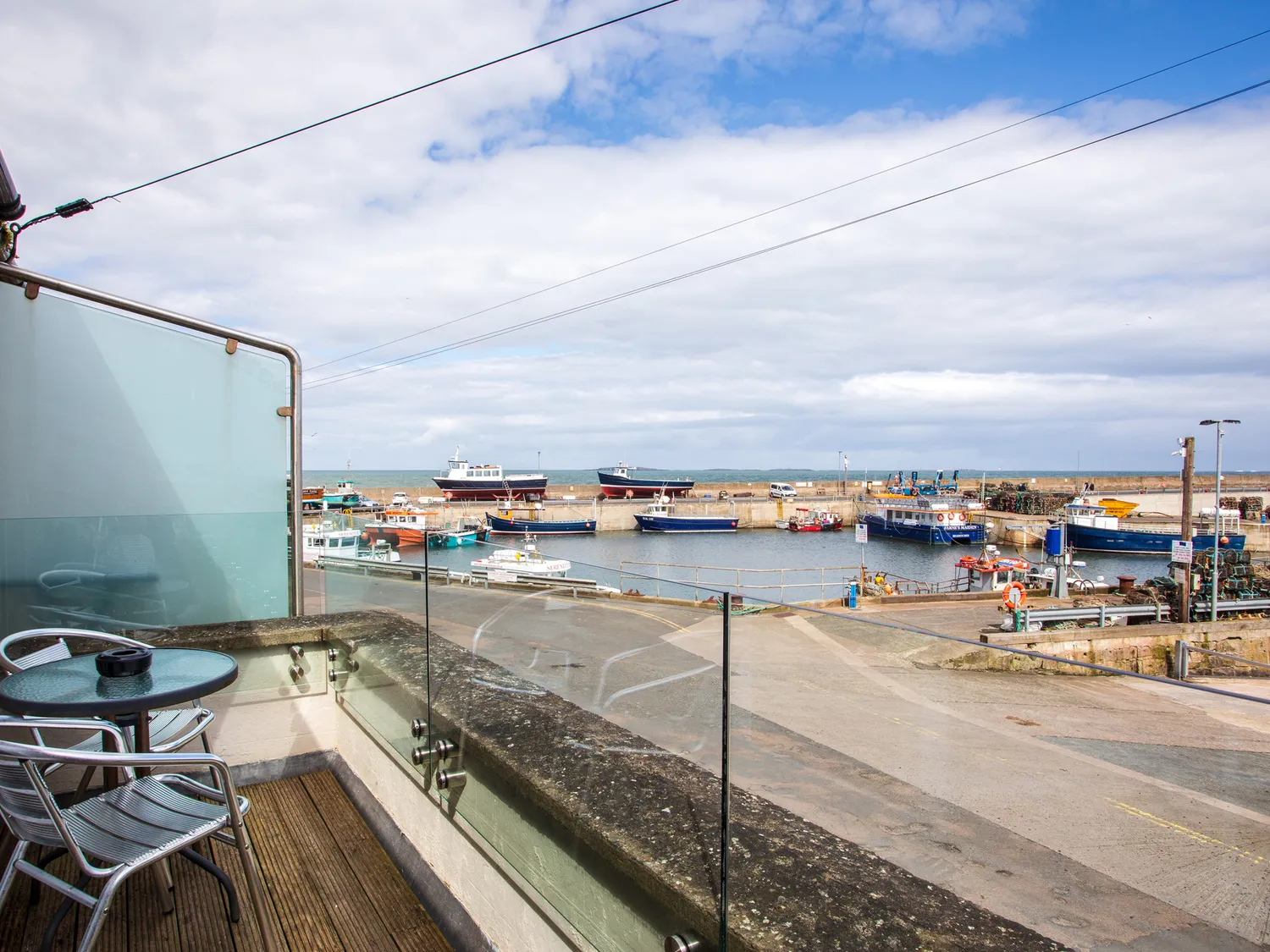 View of a marina with various boats docked, seen from a balcony with a glass railing and a small table with two chairs.