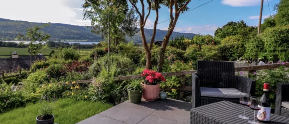 Outdoor patio with black wicker furniture, overlooking lush gardens and a scenic view of distant hills, a lake, and blue sky.