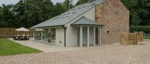 Modern country house with stone and brick exterior, large glass sliding doors, patio area with outdoor furniture, and a gravel driveway.