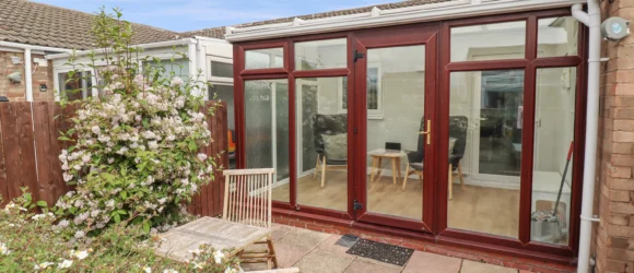 Garden patio with wooden chairs and a table, adjacent to a conservatory with large glass doors and windows framed in dark wood. Bushes with white flowers are in the foreground.