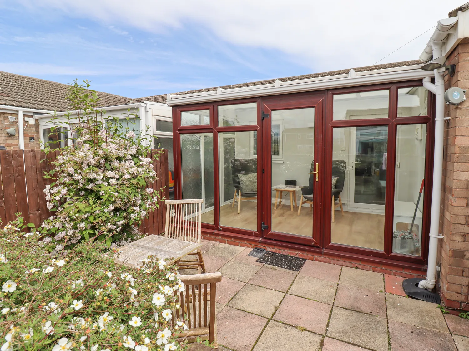 Garden patio with wooden chairs and a table, adjacent to a conservatory with large glass doors and windows framed in dark wood. Bushes with white flowers are in the foreground.