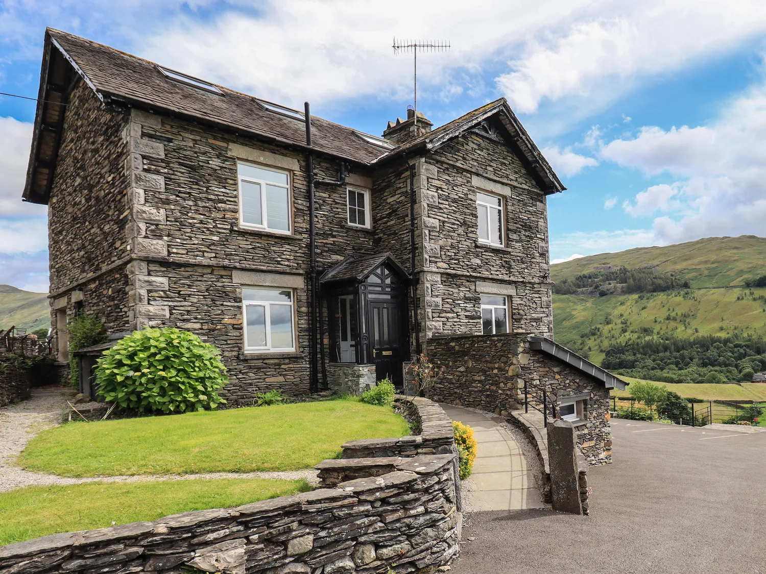 Stone-built house with a gabled roof and paved front pathway, set in a green hilly landscape under a partly cloudy sky.