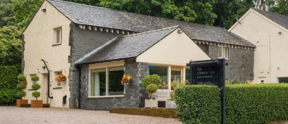 Traditional stone and stucco guest house with a gravel parking area, surrounded by lush greenery and trees.