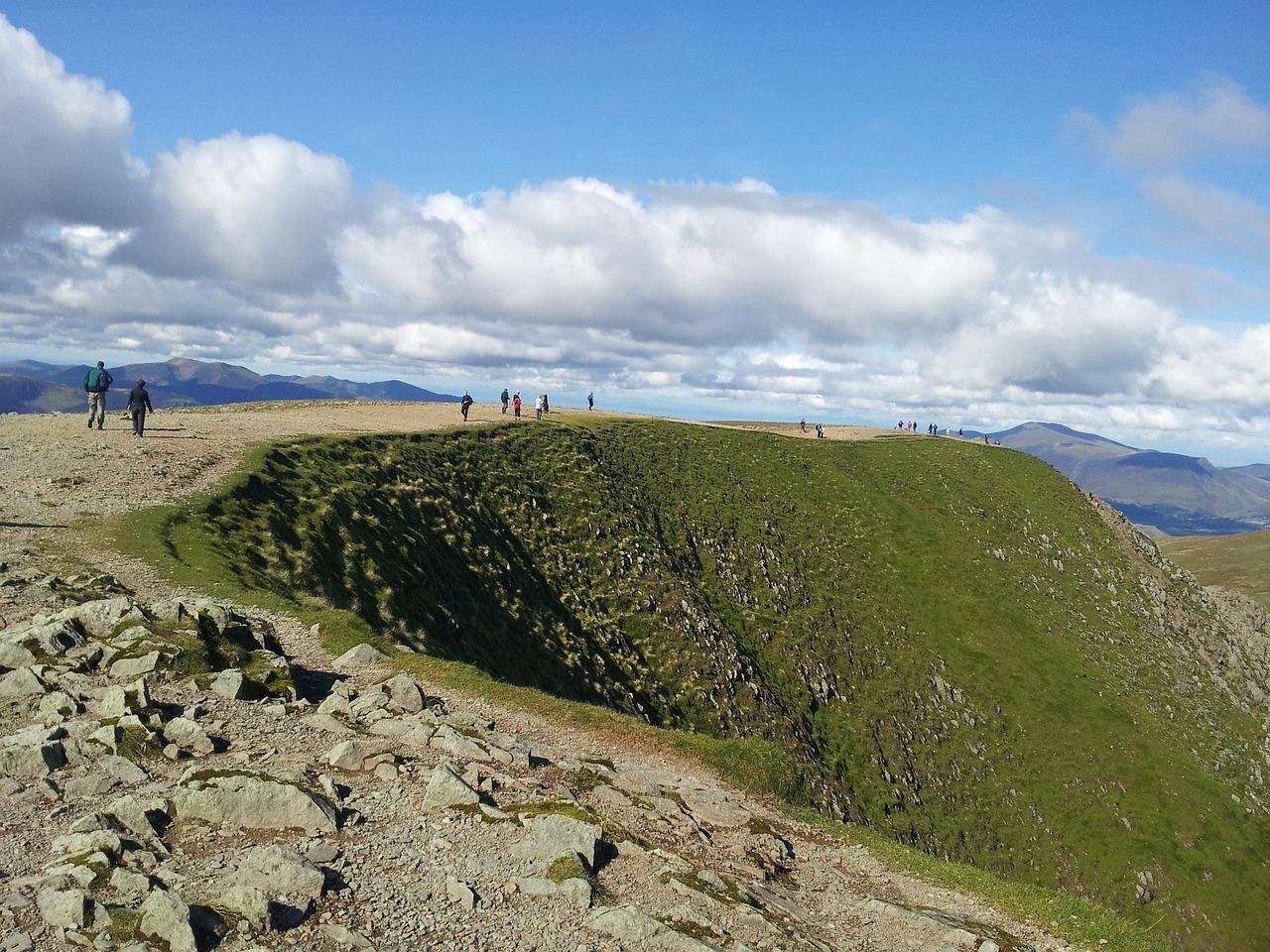 Hikers walking along a rocky and grassy ridge on a mountain under a partly cloudy sky.