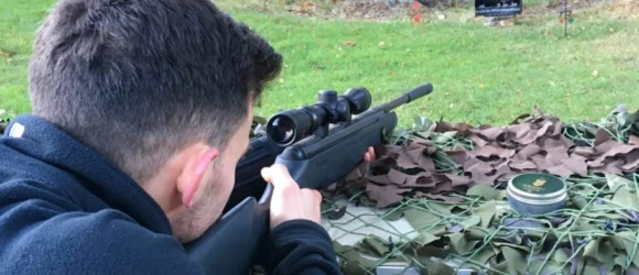 Person aiming a rifle at a target range, lying prone on the ground covered in camouflage netting.