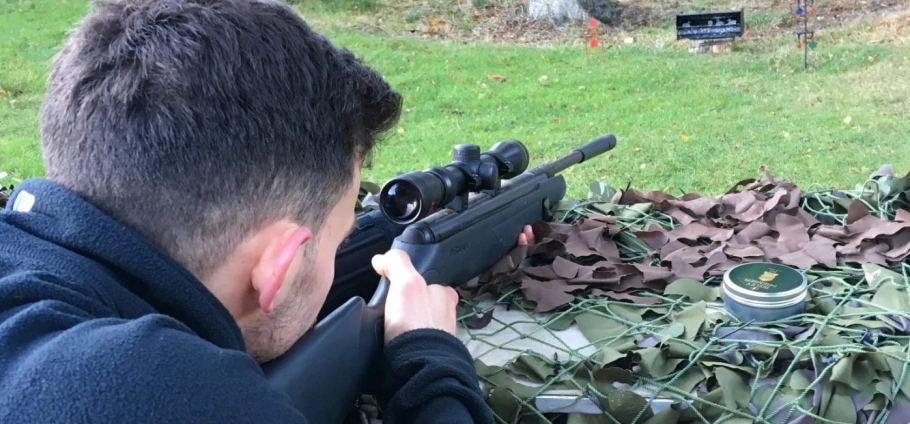 Person aiming a rifle at a target range, lying prone on the ground covered in camouflage netting.