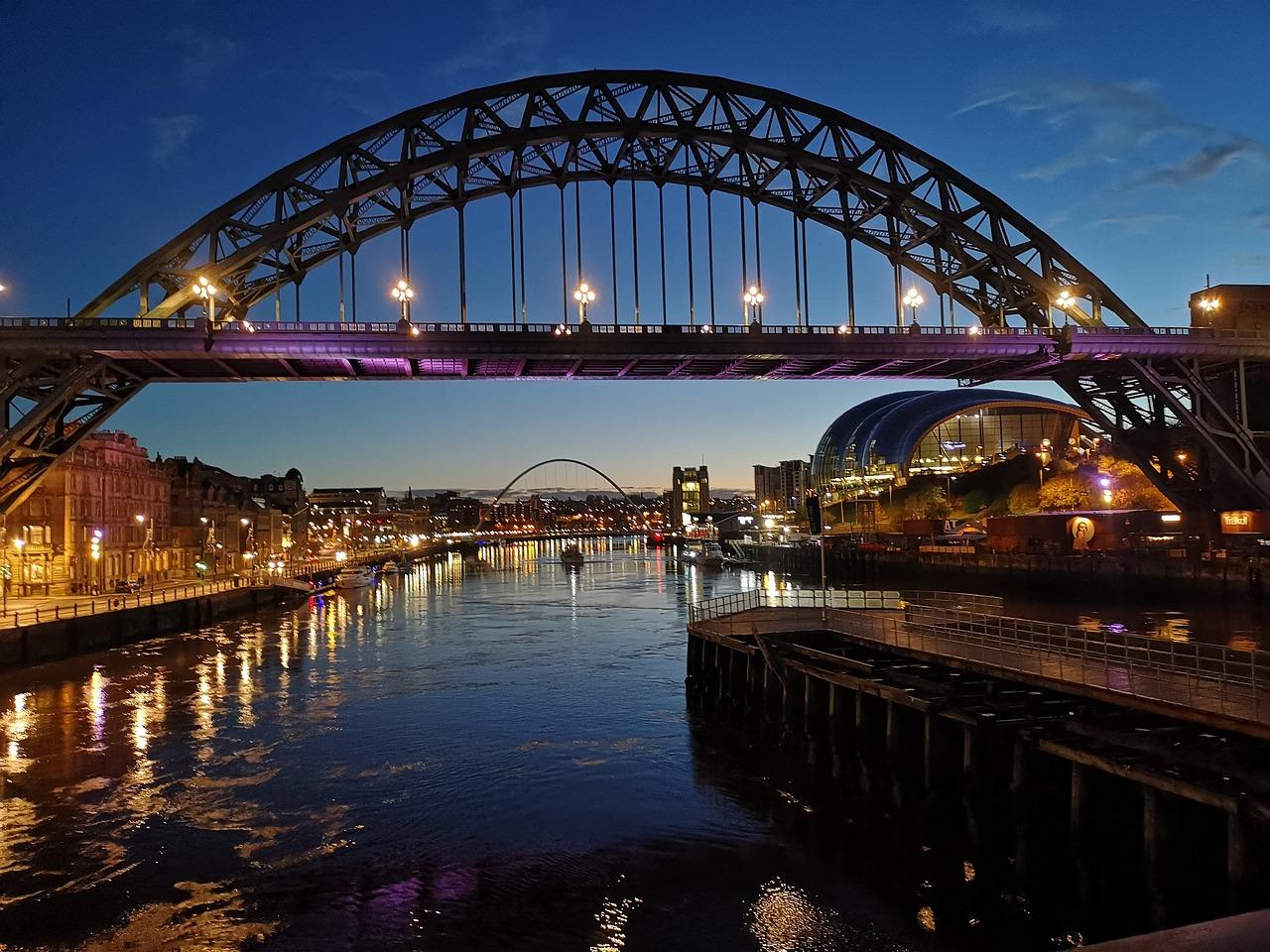 A lit-up arch bridge spanning over a river at dusk, with city lights reflecting on the water below.