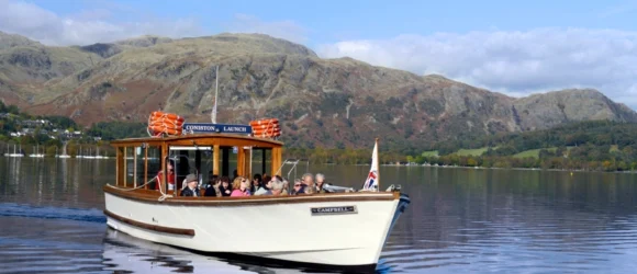 A boat carrying passengers cruises on a calm lake, with mountainous terrain and a partly cloudy sky in the background.