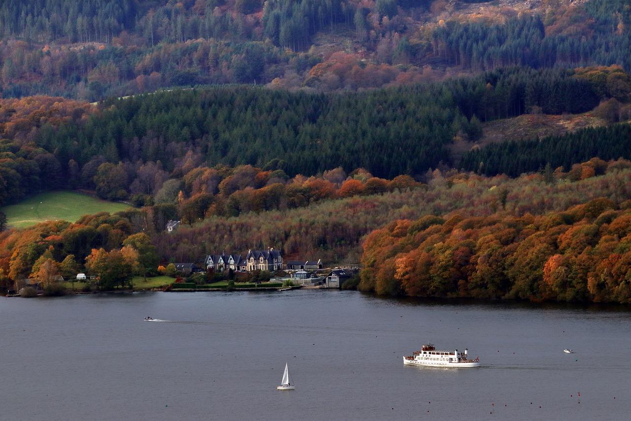 Lake Windermere | Aerial view of a lake with a sailboat and a larger boat, surrounded by autumn-coloured trees and hills. In the background, there is a village with buildings nestled among the trees.