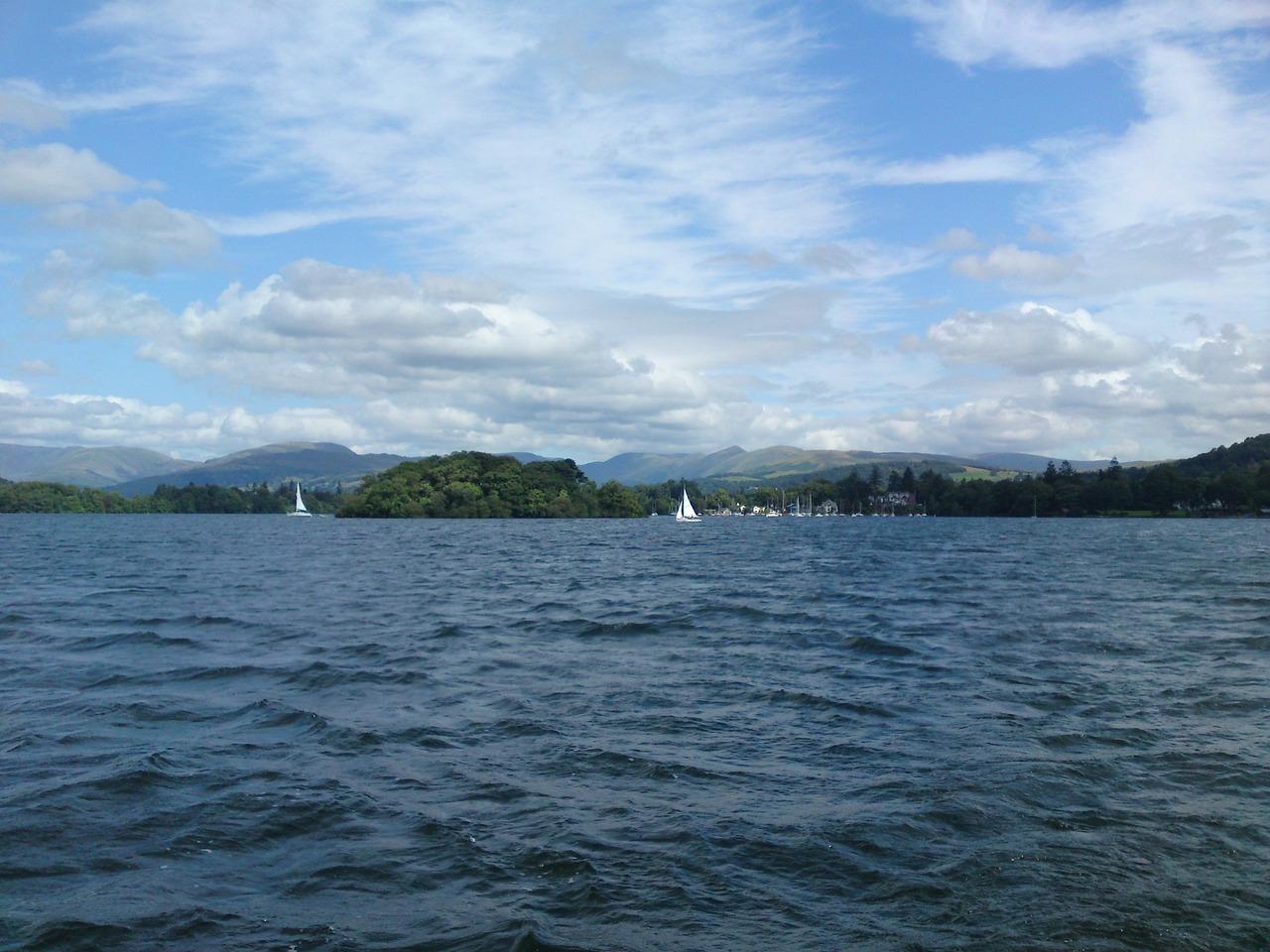 Lake with sailboats, surrounded by green hills and trees, under a partly cloudy sky.