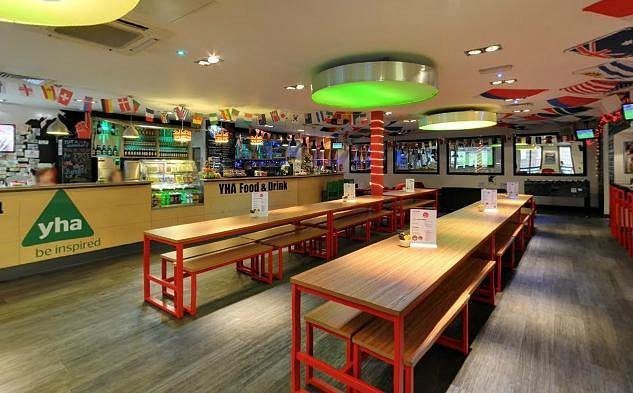 Hostel Search 1 Interior of a modern hostel with long wooden tables and benches with red metal frames, a counter in the background labeled 'YHA Food & Drink', ceiling decorated with international flags, and green circular light fixtures.